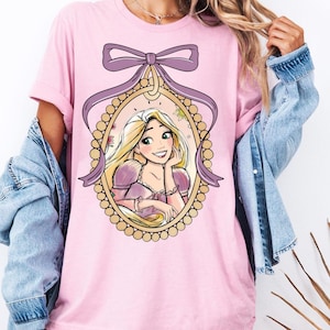 Puede incluir: Camiseta rosa claro con una ilustración de dibujos animados de una princesa en un marco ovalado dorado, adornada con un lazo y una cinta morados. La princesa tiene el pelo largo y rubio y lleva un vestido rosa. La camiseta es de estilo casual.