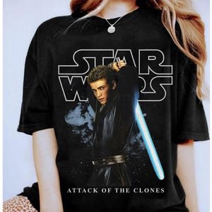 May include: Black t-shirt featuring a graphic of Anakin Skywalker from Star Wars: Attack of the Clones. The design includes the Star Wars logo in white and a blue lightsaber. The text "ATTACK OF THE CLONES" is printed at the bottom.