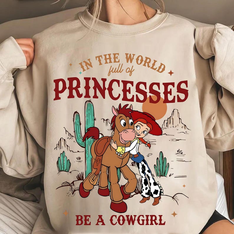 May include: Beige sweatshirt with a graphic featuring a cartoon cowgirl hugging a horse, with a desert landscape backdrop. Text reads "In the world full of princesses, be a cowgirl."