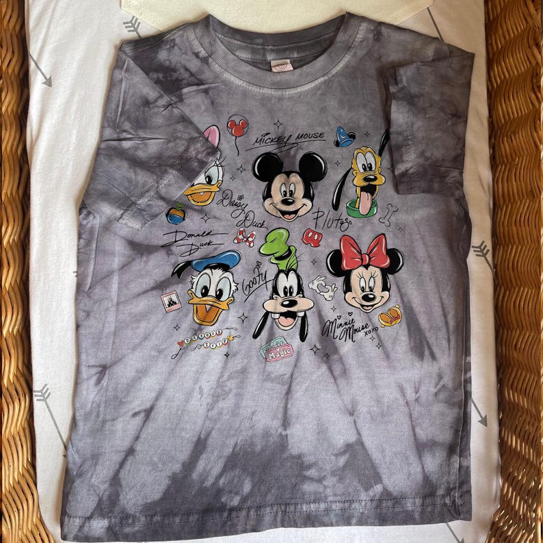 May include: Gray tie-dye t-shirt featuring Mickey Mouse, Donald Duck, Goofy, Pluto, Daisy Duck, and Minnie Mouse. The shirt has the characters' names and other graphics. The shirt is made of a soft material.