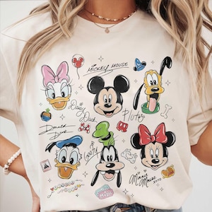 May include: Cream-colored t-shirt featuring cartoon illustrations of Disney characters Mickey Mouse, Minnie Mouse, Donald Duck, Daisy Duck, Pluto, and Goofy. The characters' names are written in cursive script. The shirt has a crew neck.