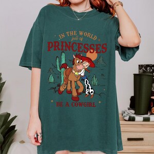 May include: Dark teal t-shirt with a graphic of a cartoon cowgirl hugging a horse, with desert scenery. Text reads "In the world full of princesses, be a cowgirl."