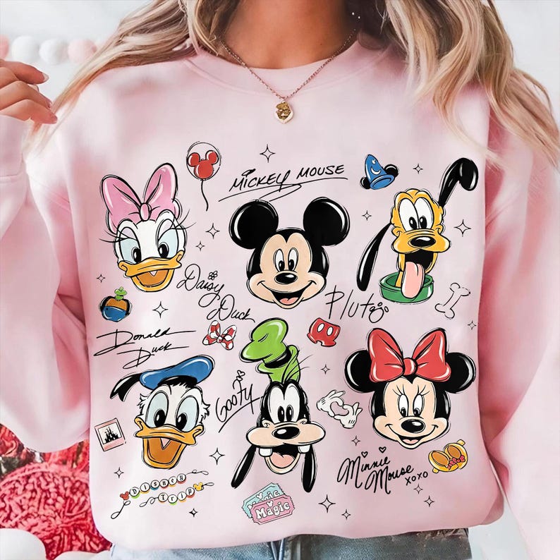 May include: A light pink sweatshirt featuring cartoon character illustrations of Mickey Mouse, Minnie Mouse, Donald Duck, Daisy Duck, Pluto, and Goofy. The characters' names are written in cursive script. The sweatshirt has a crew neck.