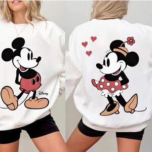 Puede incluir: Sudadera blanca con Mickey Mouse en la parte delantera y Minnie Mouse en la trasera. Mickey lleva pantalones cortos rojos y Minnie un vestido de lunares rojos. El logo de Disney es visible en la parte delantera.