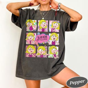 May include: Dark gray t-shirt featuring a grid of nine cartoon images of Lizzie McGuire with various expressions. The name "Lizzie McGuire" is in a pink cloud. The shirt is a relaxed fit.