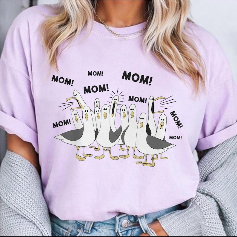 May include: A light purple t-shirt featuring a cartoon illustration of seagulls with the word "MOM!" above each bird. The seagulls are white with gray wings and yellow feet. The shirt has rolled-up sleeves.