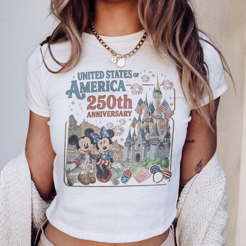 May include: Cream-colored t-shirt featuring a graphic celebrating the 250th anniversary of the United States of America. The design includes Mickey and Minnie Mouse, a castle, fireworks, and patriotic imagery.