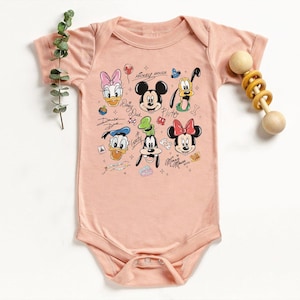 May include: A light pink baby onesie featuring a colorful graphic of Mickey Mouse and friends. The design includes Mickey, Minnie, Donald, Daisy, Goofy, and Pluto. The text "Mickey Mouse" and character names are also visible. A wooden rattle is on the right.