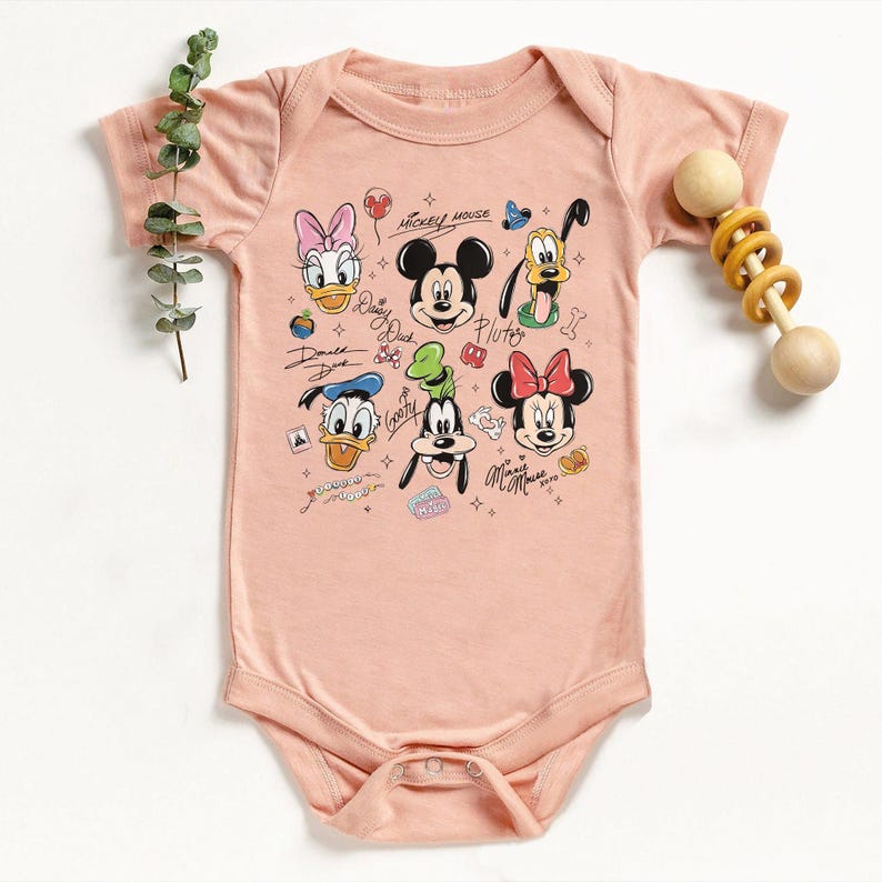 May include: A light pink baby onesie featuring a colorful graphic of Mickey Mouse and friends. The design includes Mickey, Minnie, Donald, Daisy, Goofy, and Pluto. The text "Mickey Mouse" and character names are also visible. A wooden rattle is on the right.