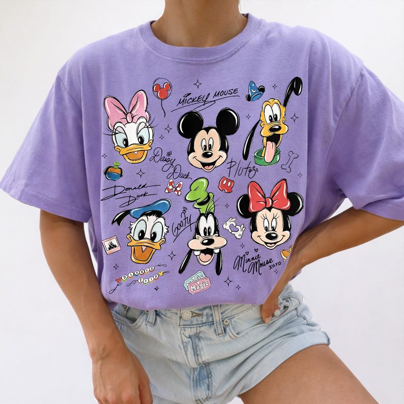 May include: A lavender t-shirt featuring a colorful graphic of Disney characters, including Mickey Mouse, Minnie Mouse, Donald Duck, Daisy Duck, Goofy, and Pluto. The design includes character signatures and other small illustrations.