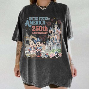 May include: Dark gray t-shirt featuring a graphic commemorating the 250th anniversary of the United States of America. The design includes Mickey and Minnie Mouse, a castle, and patriotic imagery.