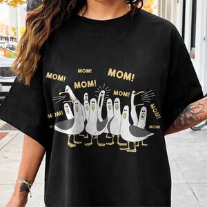 May include: Black t-shirt featuring a cartoon illustration of seagulls with the word "MOM!" in gold lettering. The seagulls are in various poses, with some squawking and others looking surprised. The shirt is made of a soft material.
