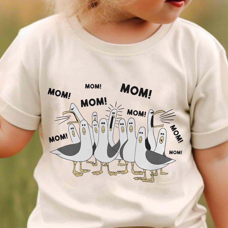 May include: A light beige t-shirt featuring a cartoon illustration of seagulls with the word "MOM!" above each bird. The seagulls are white with gray wings and yellow feet. The shirt is made of a soft material.