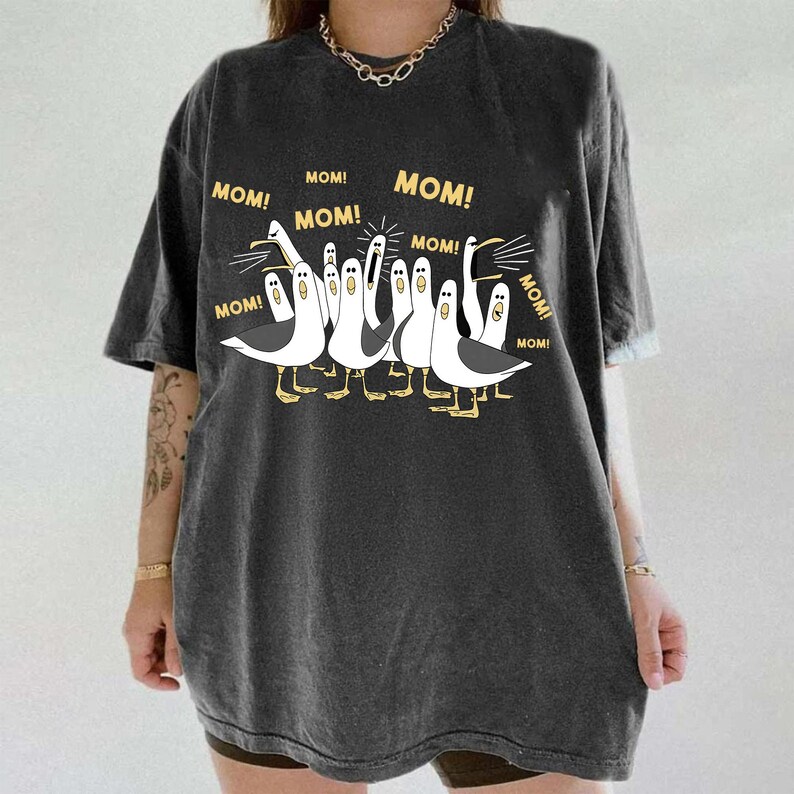 May include: Dark gray t-shirt featuring a cartoon illustration of seagulls with the word "MOM!" above each bird. The seagulls are white with black accents and yellow feet. The shirt has short sleeves and a relaxed fit.