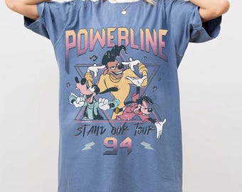 Op de afbeelding: Blauw T-shirt met het woord "POWERLINE" en een afbeelding van drie stripfiguren. De tekst "STAND OUT TOUR 94" staat eronder. Het shirt heeft een ronde hals en korte mouwen.