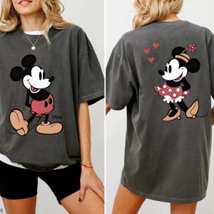 Puede incluir: Camiseta gris oscuro con Mickey Mouse en la parte delantera y Minnie Mouse en la parte trasera. Mickey est&aacute; en una pose cl&aacute;sica con pantalones cortos rojos. Minnie lleva un vestido de lunares y una flor. El logotipo de Disney es visible.