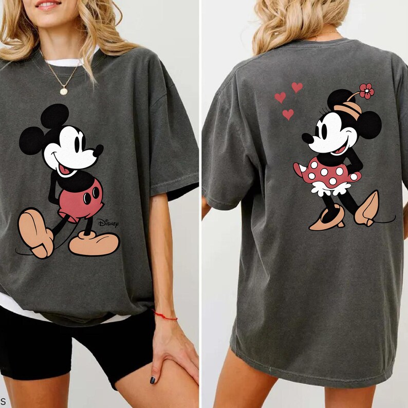 Puede incluir: Camiseta gris oscuro con Mickey Mouse en la parte delantera y Minnie Mouse en la parte trasera. Mickey est&aacute; en una pose cl&aacute;sica con pantalones cortos rojos. Minnie lleva un vestido de lunares y una flor. El logotipo de Disney es visible.