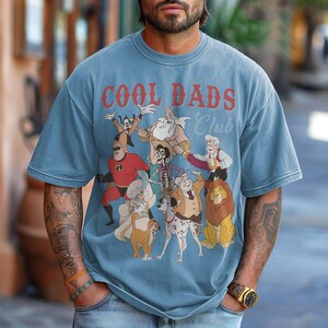 K&ouml;nnte beinhalten: Hellblaues T-Shirt mit einem Aufdruck von Zeichentrickfiguren und dem Text "COOL DADS CLUB" in Rot. Das Design umfasst Charaktere wie Mr. Incredible und Scar. Das T-Shirt hat einen Vintage-Look.