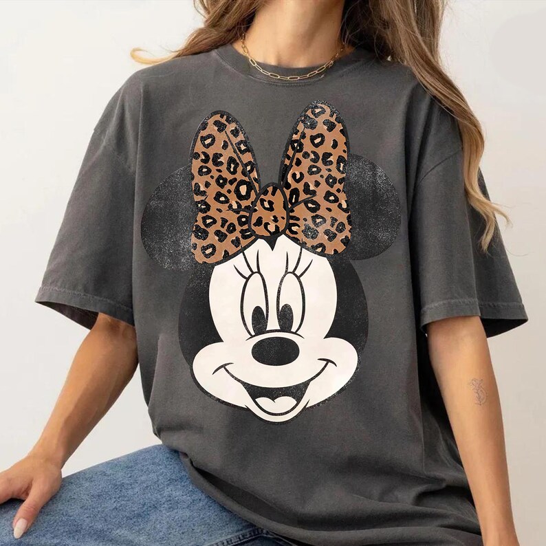 K&ouml;nnte beinhalten: Dunkelgraues T-Shirt mit einem Minnie Mouse-Motiv. Die Grafik zeigt ein schwarz-wei&szlig;es Minnie Mouse-Gesicht mit einer Schleife im Leopardenmuster. Das T-Shirt hat kurze &Auml;rmel und eine lockere Passform, geeignet f&uuml;r den t&auml;glichen Gebrauch.