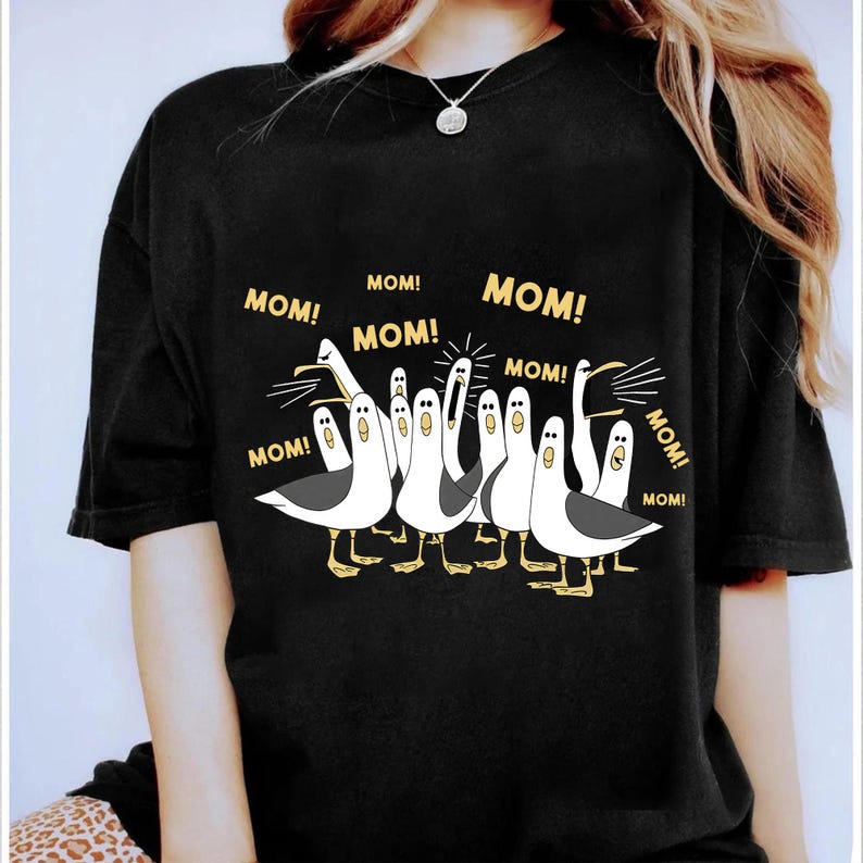 May include: Black t-shirt featuring a cartoon illustration of seagulls with the word "MOM!" repeated around them. The seagulls are white with black wings and yellow feet. The shirt is a casual, comfortable style.