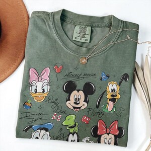 May include: Green Comfort Colors t-shirt featuring Mickey Mouse and friends. The shirt has a crew neck and short sleeves. The graphic includes Mickey, Minnie, Donald, Daisy, Pluto, and Goofy. A gold necklace is draped across the shirt.