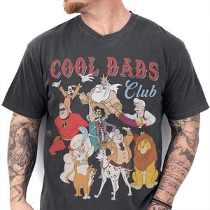 Puede incluir: Camiseta gris oscuro con cuello en V con el texto "COOL DADS Club" en rojo y azul. La camiseta presenta un gráfico colorido de personajes de dibujos animados, incluyendo Mickey Mouse, Mr. Incredible y Simba.