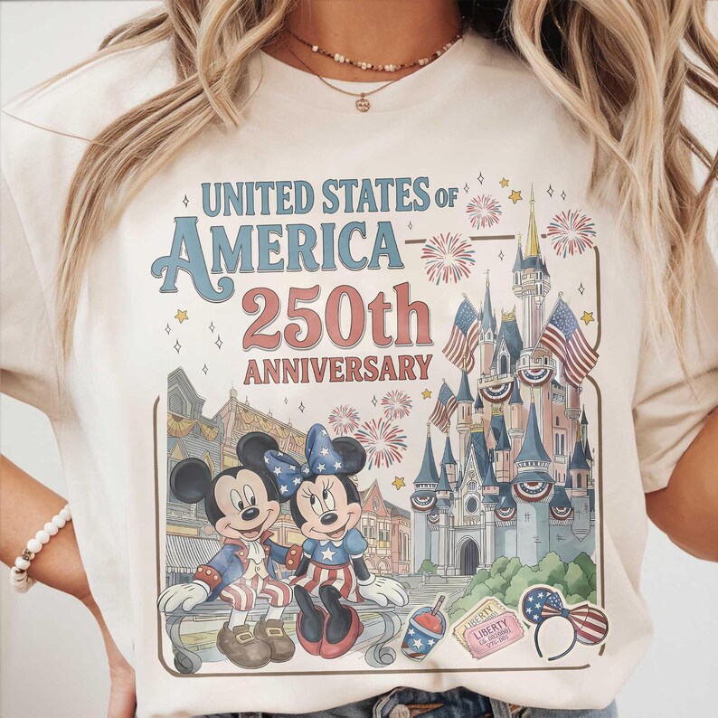 May include: Cream-colored t-shirt featuring a graphic celebrating the 250th anniversary of the United States of America. The design includes a castle, fireworks, and Mickey and Minnie Mouse in patriotic attire.