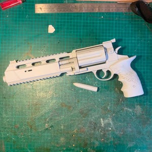 May include: A matte white 3D printed revolver prop with a long barrel, a top rail, and a cylinder. The grip features an engraved wing design. A single white bullet-shaped component lies beneath the barrel, next to a metal ruler showing 28 cm.