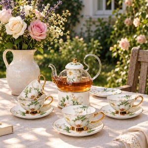 Peut inclure: Une théière en verre avec un motif floral et un chauffe-théière assorti, accompagnée de trois tasses en porcelaine et soucoupes à motifs de fleurs blanches et anses dorées, disposées sur une table avec un pichet en céramique de roses.