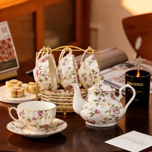 May include: A white porcelain tea set featuring a delicate pink rose and green vine pattern with gold-trimmed edges. The set includes a teapot, multiple teacups with matching saucers, and a gold-toned metal rack for holding the cups and saucers.