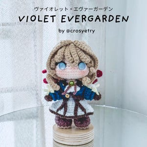 Puede incluir: Una muñeca de ganchillo hecha a mano de Violet Evergarden con cabello castaño claro, ojos azules y un atuendo azul y blanco. La muñeca presenta una cuenta verde en el cuello y detalles rojos. El texto superior dice: ヴァイオレット・エヴァーガーデン VIOLET EVERGARDEN by @crosyetry.