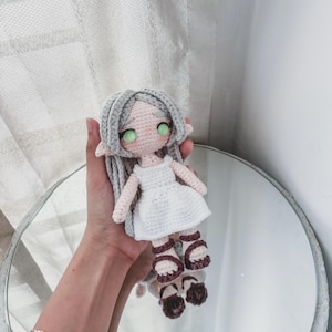 May include: A handmade crochet doll with grey hair, green eyes, and a white dress. The doll is wearing brown sandals. The doll is being held in front of a mirror.