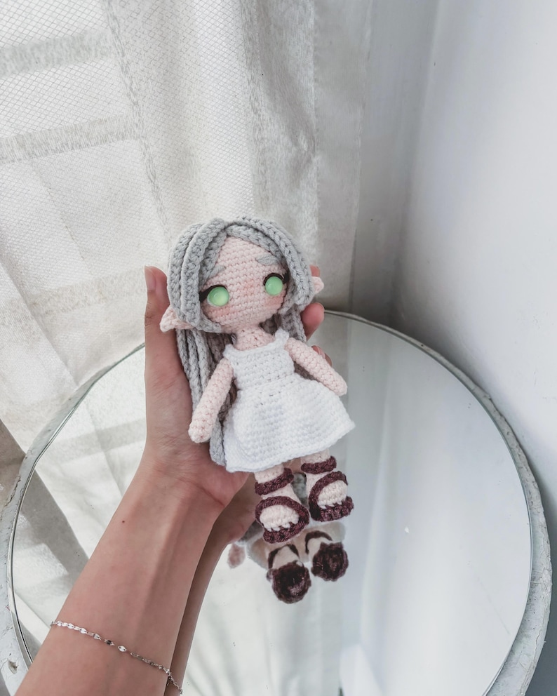 May include: A handmade crochet doll with grey hair, green eyes, and a white dress. The doll is wearing brown sandals. The doll is being held in front of a mirror.