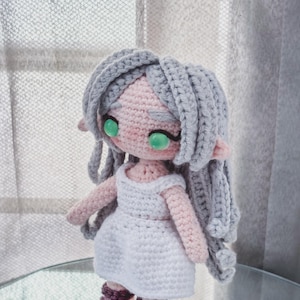 May include: A handmade crochet doll with grey hair, pink skin, and green eyes. The doll wears a white dress and purple and white sandals. The doll has long grey hair and is standing on a glass surface.