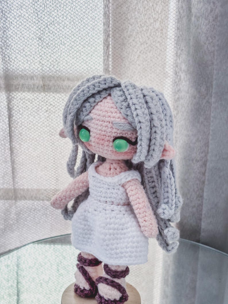 May include: A handmade crochet doll with grey hair, pink skin, and green eyes. The doll wears a white dress and purple and white sandals. The doll has long grey hair and is standing on a glass surface.