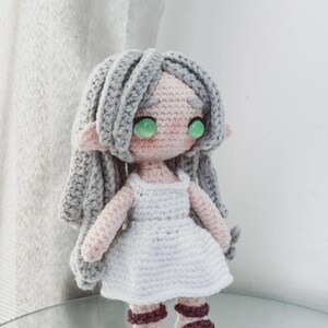 May include: A handcrafted crochet doll with grey hair, green eyes, and a pink face. The doll is dressed in a white dress and burgundy sandals. The doll has pointed ears, suggesting an elven or fairy theme.