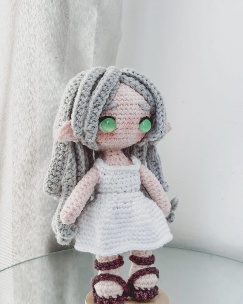 May include: A handcrafted crochet doll with grey hair, green eyes, and a pink face. The doll is dressed in a white dress and burgundy sandals. The doll has pointed ears, suggesting an elven or fairy theme.