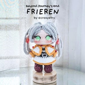 Op de afbeelding: Een gehaakte pop van het personage Frieren uit Beyond Journey's End, met lichtgrijs haar, grote groene ogen, een wit en goudkleurig outfit en donkerrode laarzen. De tekst Beyond Journey's End FRIEREN by @crosyetry is zichtbaar.