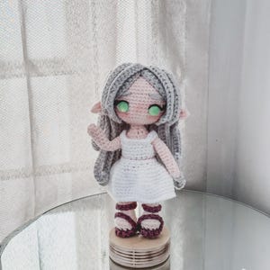 May include: A handmade crochet doll with grey hair, green eyes, and a white dress. The doll is wearing burgundy sandals and is standing on a wooden platform. The doll has a sweet, whimsical appearance.
