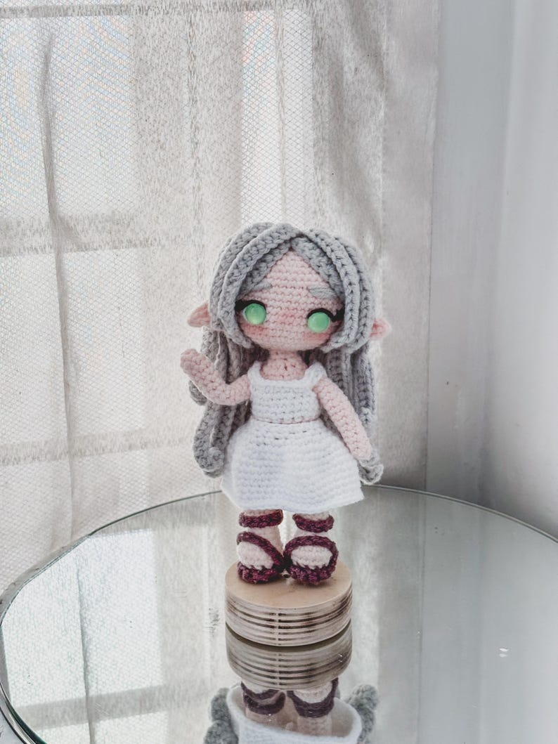 May include: A handmade crochet doll with grey hair, green eyes, and a white dress. The doll is wearing burgundy sandals and is standing on a wooden platform. The doll has a sweet, whimsical appearance.