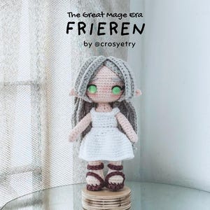 May include: Crocheted doll of a character named Frieren, with grey hair, green eyes, and a white dress. The doll is standing on a wooden base. Text on the image reads "The Great Mage Era FRIEREN by @crosyetry."