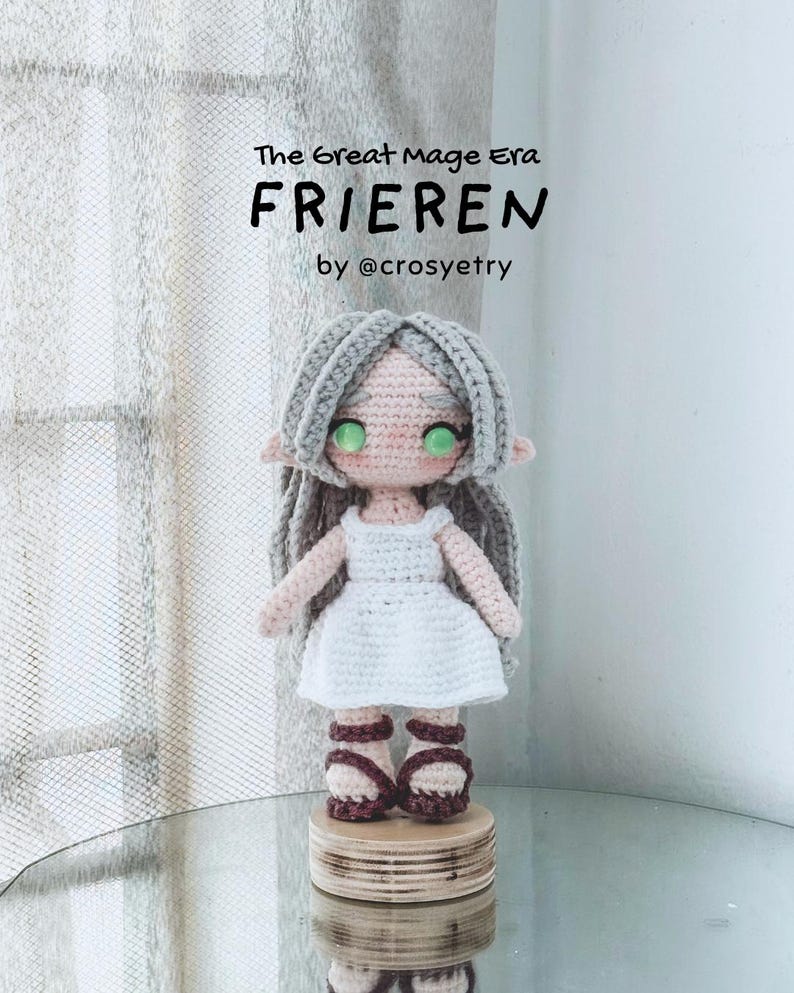 May include: Crocheted doll of a character named Frieren, with grey hair, green eyes, and a white dress. The doll is standing on a wooden base. Text on the image reads "The Great Mage Era FRIEREN by @crosyetry."
