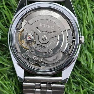 May include: A silver-toned stainless steel wristwatch with an exposed mechanical movement visible through the clear case back. The watch features a metal link bracelet and a circular case with the Seiko brand name engraved on the internal rotor.