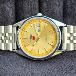 May include: A Seiko 5 automatic wristwatch with a gold-toned dial featuring horizontal lines, silver-colored hands and hour markers, and a day-date window displaying SAT 16. The watch has a stainless steel case and a matching link bracelet.