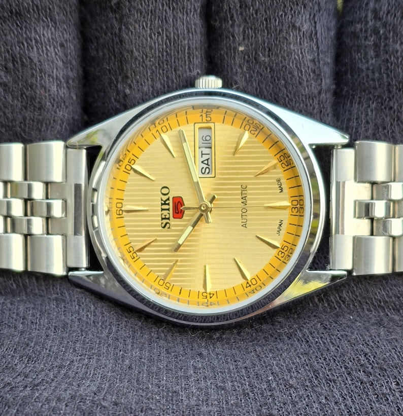 May include: A Seiko 5 automatic wristwatch with a gold-toned dial featuring horizontal lines, silver-colored hands and hour markers, and a day-date window displaying SAT 16. The watch has a stainless steel case and a matching link bracelet.