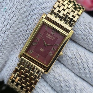 Könnte beinhalten: Eine rechteckige, goldfarbene Seiko Quartz Armbanduhr mit einem strukturierten burgunderroten Zifferblatt und römischen Ziffern. Die Uhr verfügt über ein passendes, poliertes Gliederarmband aus goldfarbenem Metall und eine kleine Krone an der Seite.