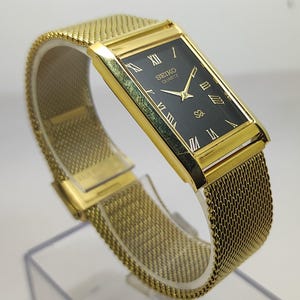 Peut inclure: Une montre Seiko Quartz dor&eacute;e avec un cadran noir rectangulaire et des chiffres romains dor&eacute;s. La montre est dot&eacute;e d'un bracelet en maille et est pr&eacute;sent&eacute;e sur un support transparent. Le cadran porte le logo Seiko Quartz.
