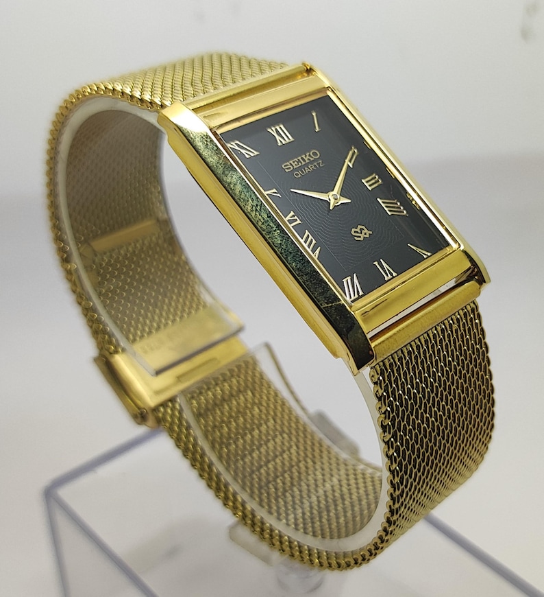 Peut inclure: Une montre Seiko Quartz dor&eacute;e avec un cadran noir rectangulaire et des chiffres romains dor&eacute;s. La montre est dot&eacute;e d'un bracelet en maille et est pr&eacute;sent&eacute;e sur un support transparent. Le cadran porte le logo Seiko Quartz.