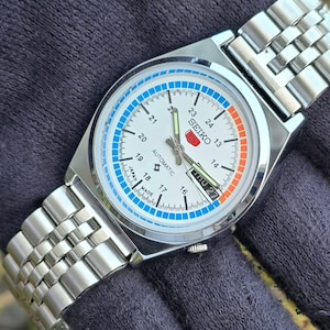 May include: A silver-toned Seiko automatic wristwatch with a white face. The watch features blue and orange accents, a day-date display, and a stainless steel band. The watch face displays the text "SEIKO" and "AUTOMATIC".