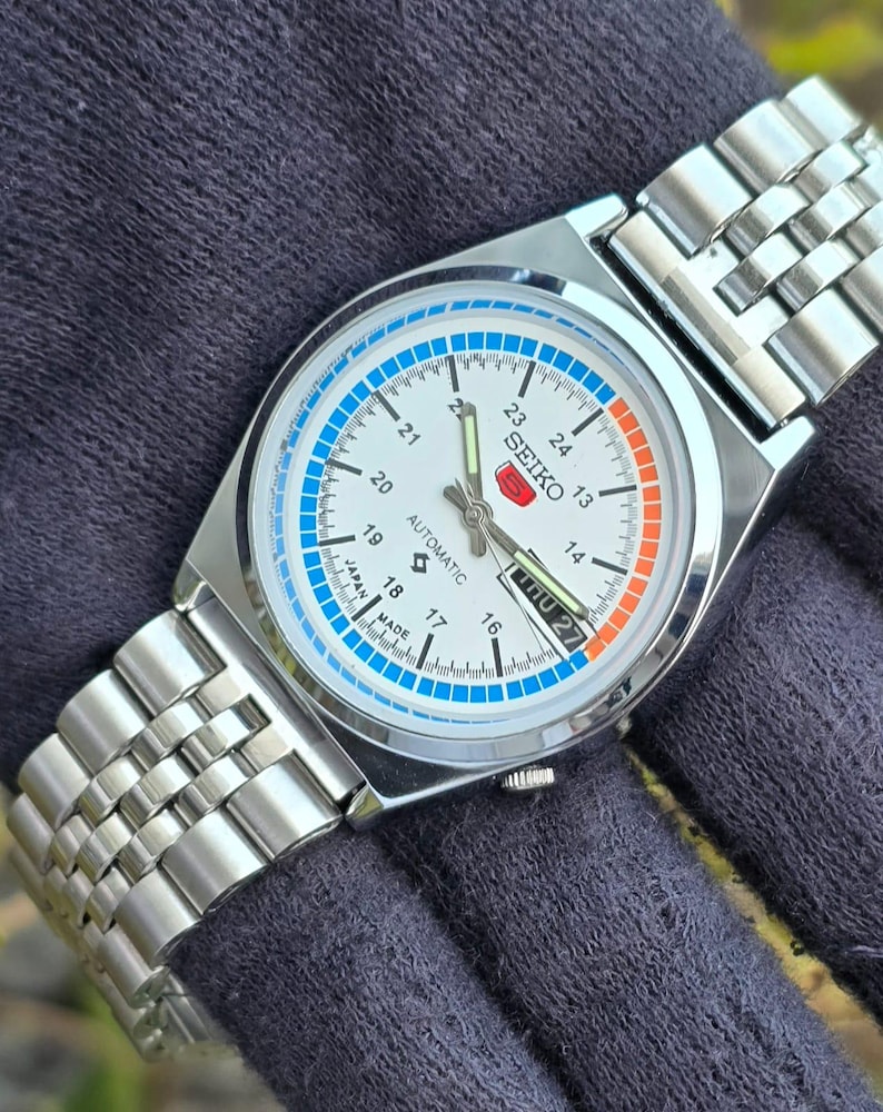 May include: A silver-toned Seiko automatic wristwatch with a white face. The watch features blue and orange accents, a day-date display, and a stainless steel band. The watch face displays the text "SEIKO" and "AUTOMATIC".